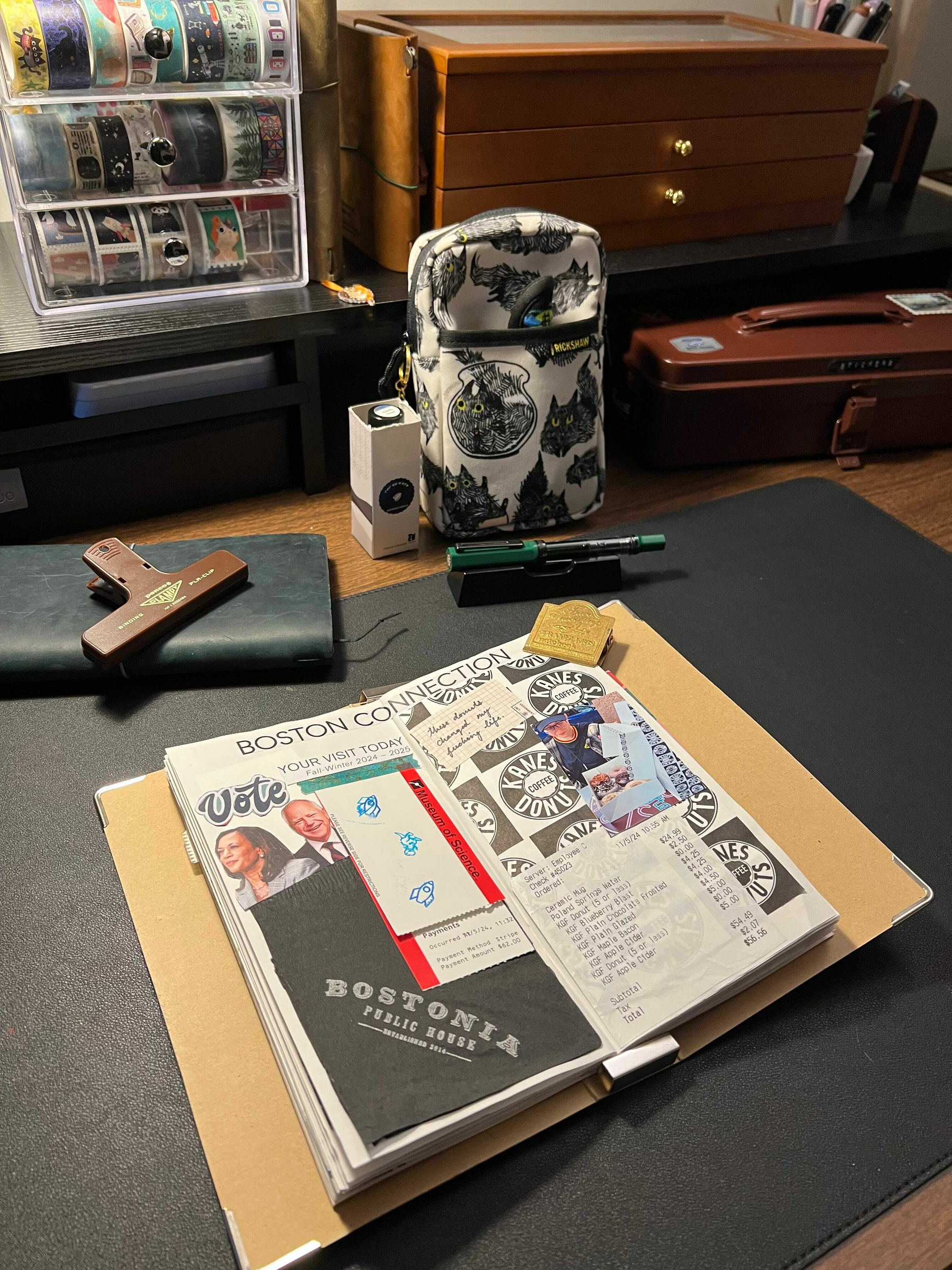Auto-generated description: A journal is open on a desk surrounded by various items, including a cat-themed pouch, a small suitcase, and decorative storage boxes.